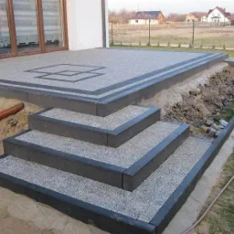 Wykończony taras z kostki brukowej z czterema stopniami, obrzeża z ciemnej kostki, w tle dom jednorodzinny i pole.