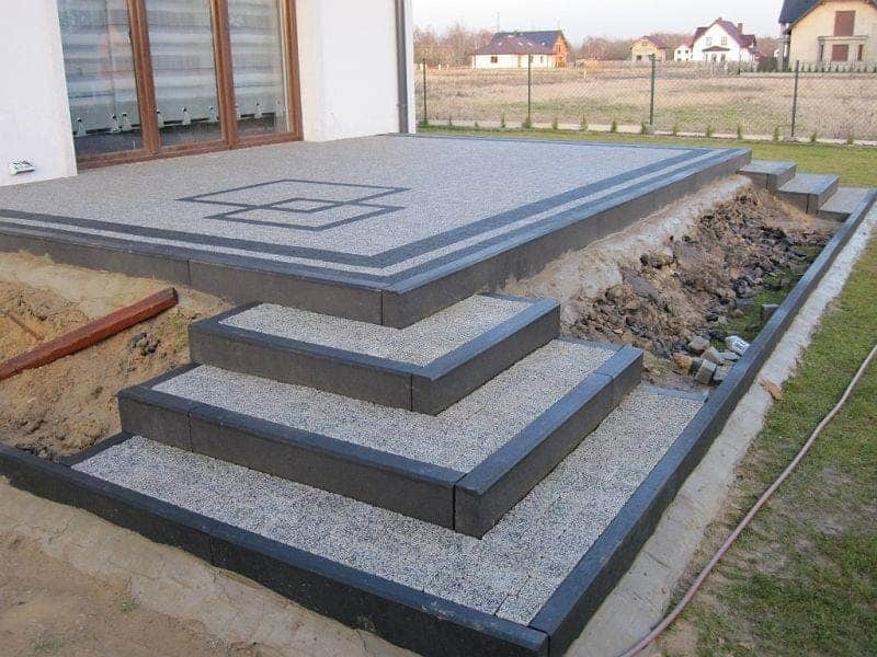 Wykończony taras z kostki brukowej z czterema stopniami, obrzeża z ciemnej kostki, w tle dom jednorodzinny i pole.