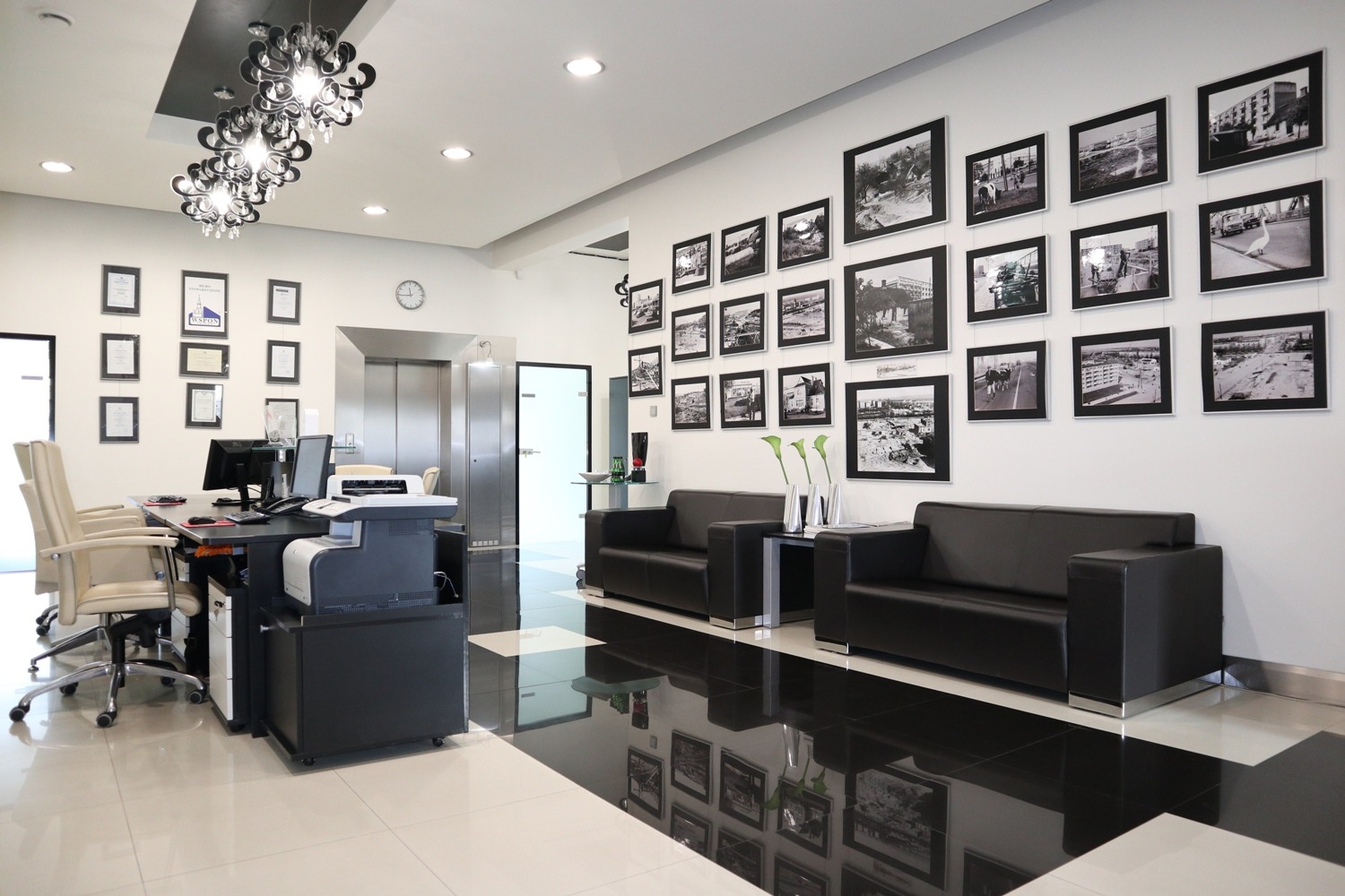 Jasne, przestronne biuro agencji nieruchomości z czarnymi sofami, biurkiem z komputerem, windą, galerią czarno-białych zdjęć na ścianie i designerskim żyrandolem.