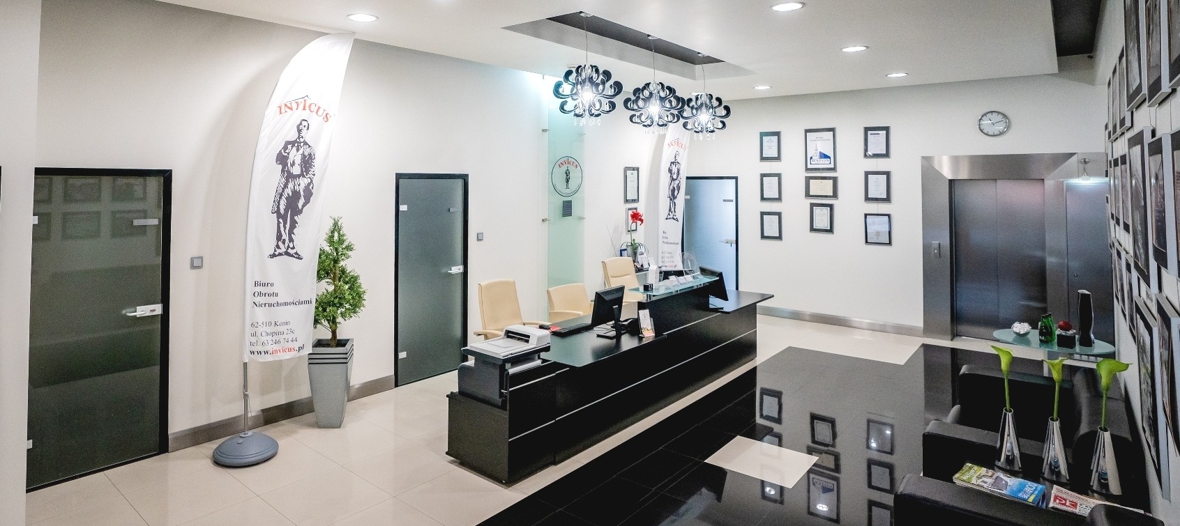 Wnętrze biura nieruchomości z recepcją, flagą z logo firmy Invictus, windą i galerią zdjęć na ścianie.