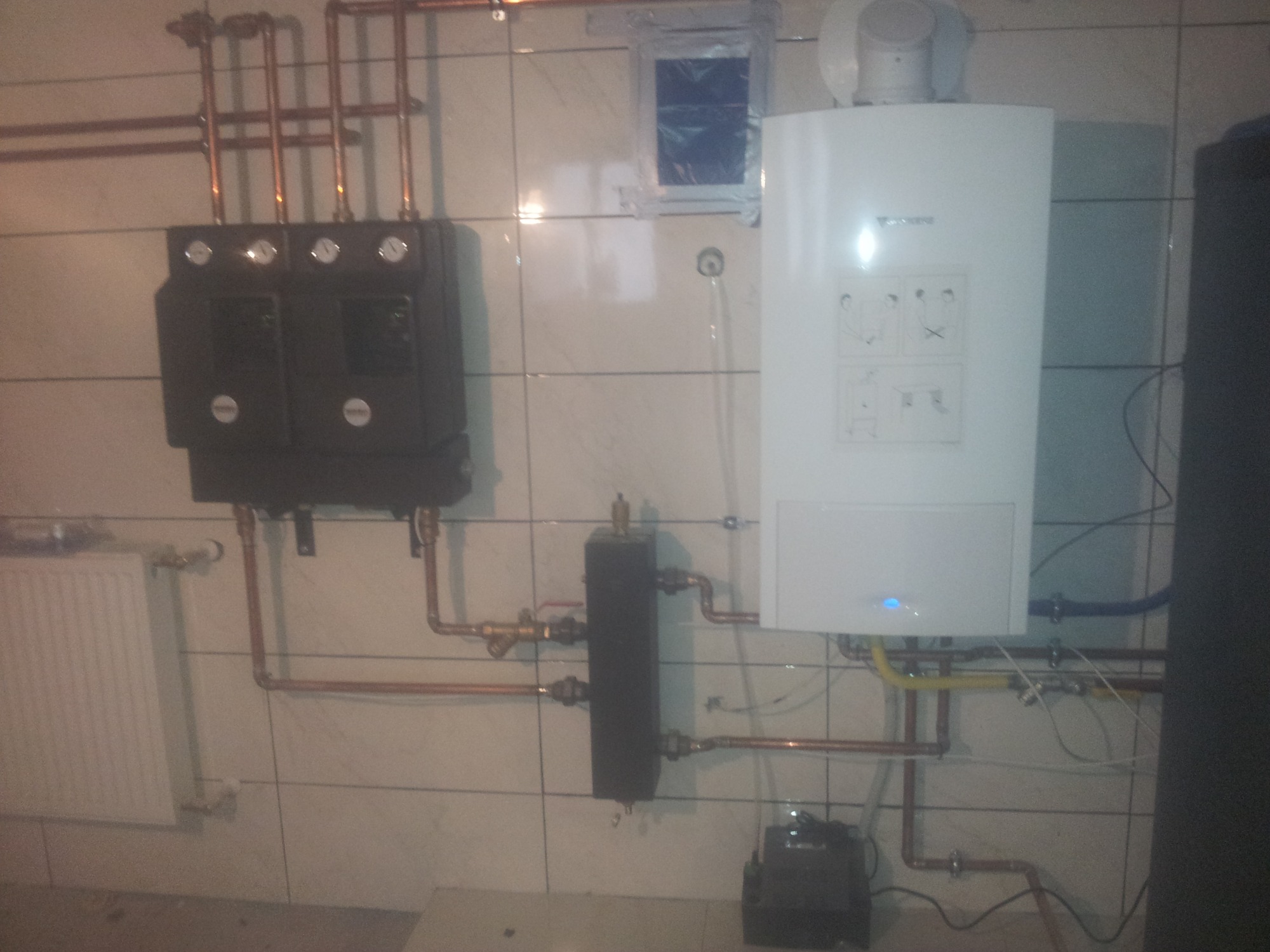 Nowoczesny piec centralnego ogrzewania marki Junkers, ciemny rozdzielacz z manometrami, miedziane rury instalacji grzewczej na tle jasnych płytek ceramicznych w kotłowni.