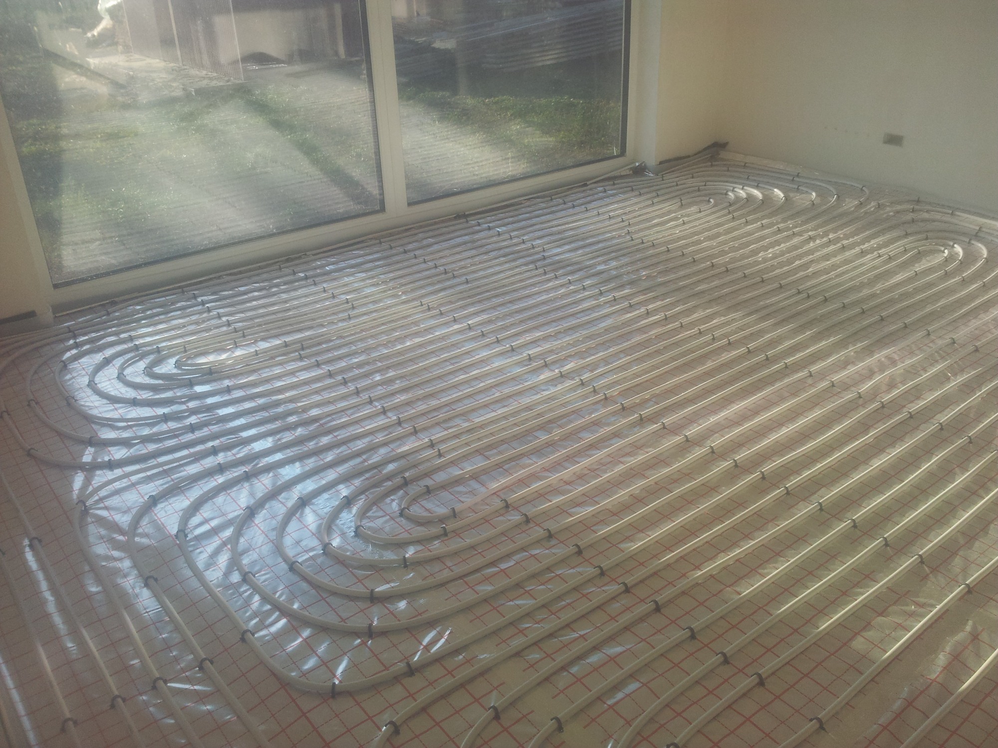 Rozłożona instalacja ogrzewania podłogowego z rur PEX na folii z nadrukowaną siatką, widok z góry, duże okno w tle