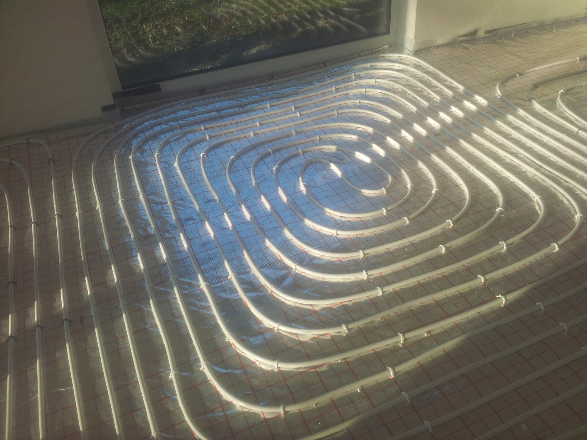 Rozłożona instalacja ogrzewania podłogowego z białych rurek na folii aluminiowej z siatką, widok z góry, częściowo odbite okno.