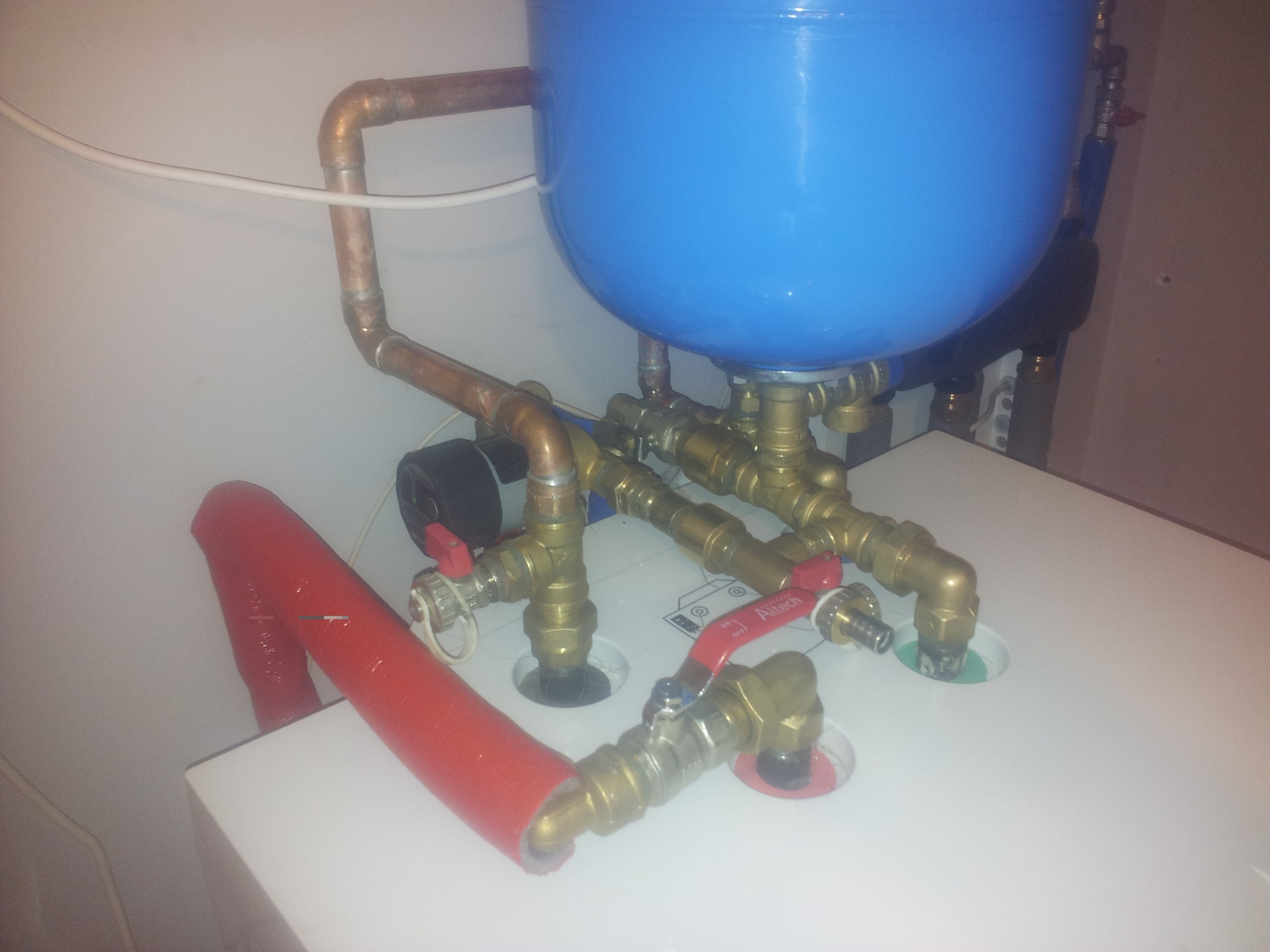 Niebieski zbiornik wyrównawczy z miedzianymi rurami i zaworami, widoczna izolacja termiczna rury w kolorze czerwonym, instalacja grzewcza w kotłowni, ujęcie z góry pod kątem.