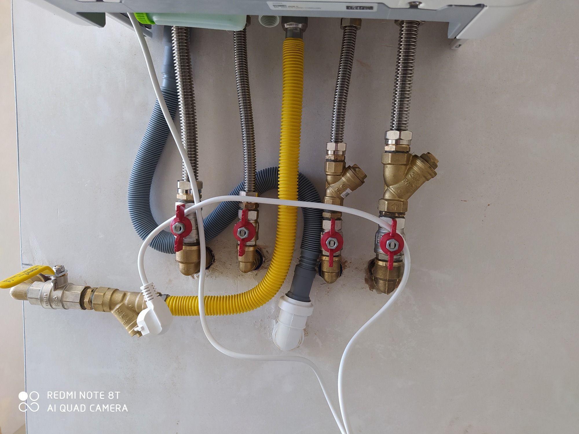 Podłączenia do pieca gazowego: elastyczne węże w oplocie stalowym, rury gazowe z zaworami odcinającymi z czerwonymi pokrętłami, szary karbowany wąż odprowadzający skropliny, biały przewód...