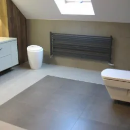 Nowoczesna łazienka na poddaszu ze skośnym sufitem i oknem dachowym, szare płytki podłogowe z wydzieloną strefą, biała umywalka z szafką, toaleta podwieszana i grafitowy grzejnik drabinkowy.