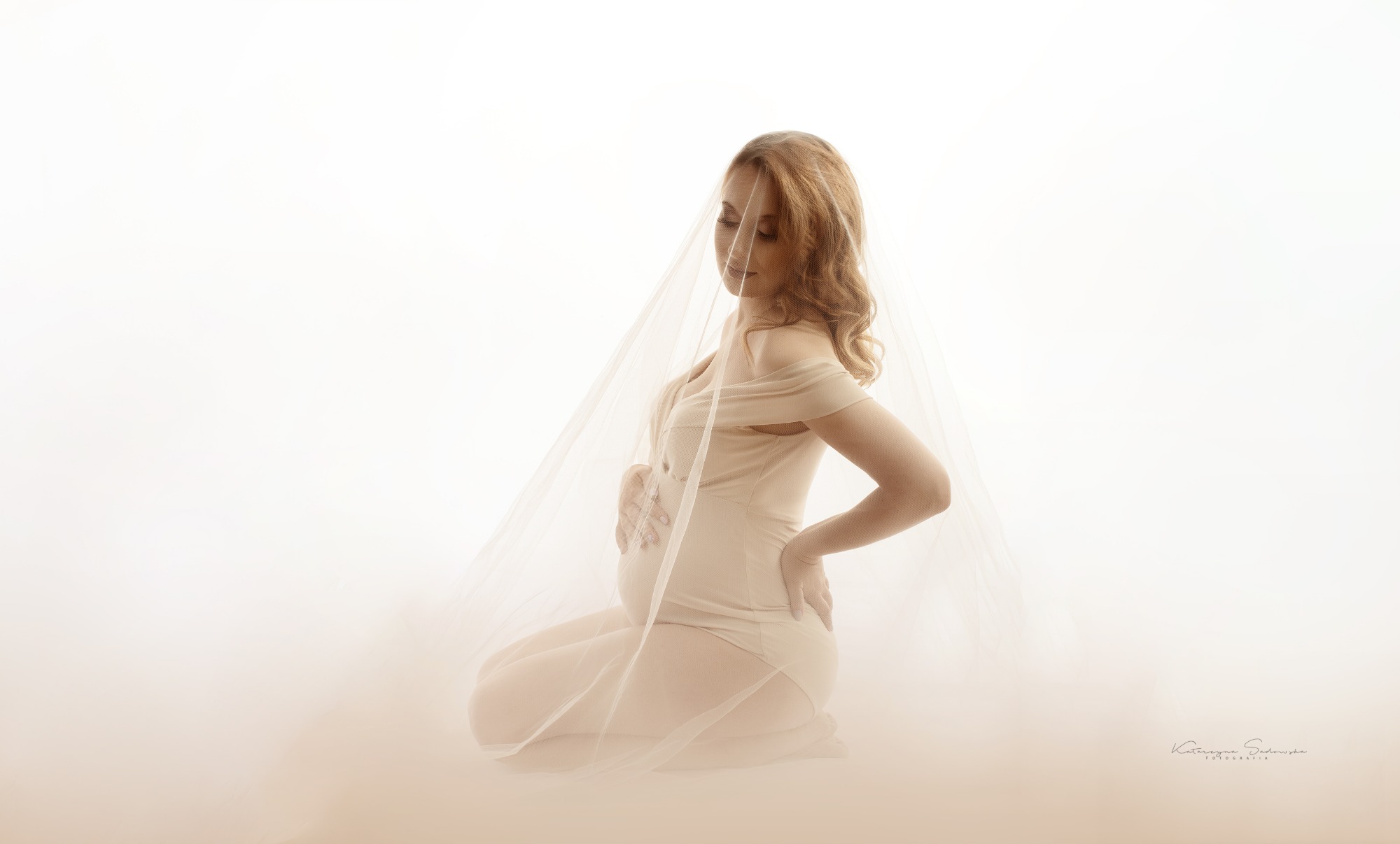 Kobieta w beżowej sukni, klęcząca, z rękami na brzuchu, otulona delikatnym woalem na jasnym tle.