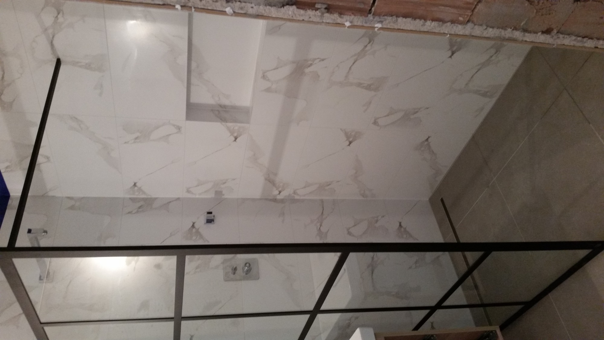 Szklana kabina prysznicowa z czarnymi profilami, zamontowana w łazience wyłożonej białymi płytkami imitującymi marmur, widoczne elementy instalacji wodnej i fragment surowego sufitu.