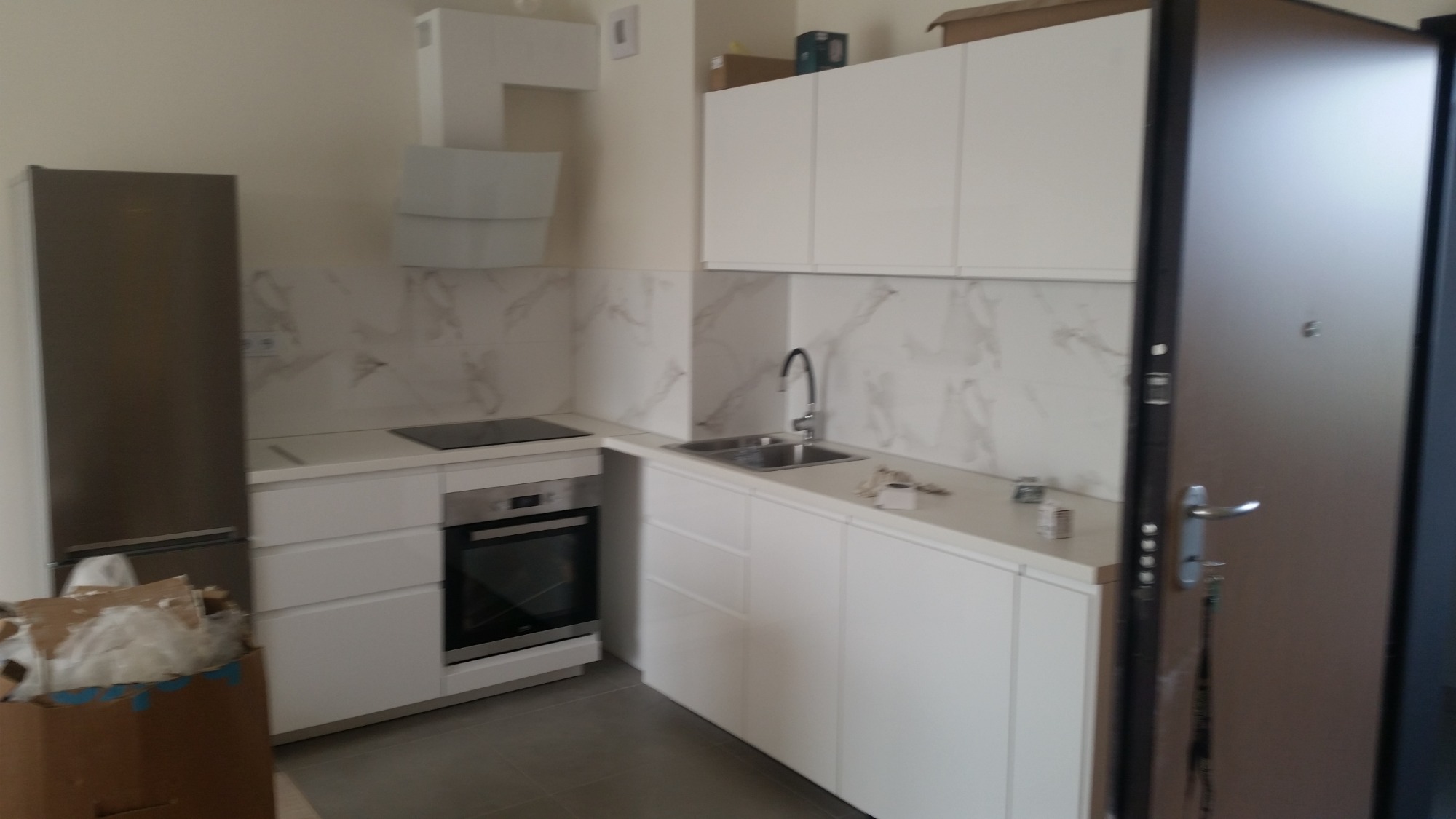 Nowoczesna, jasna kuchnia z białymi szafkami, marmurowymi płytkami na ścianie i srebrną lodówką, częściowo zasłonięta kartonami po remoncie.