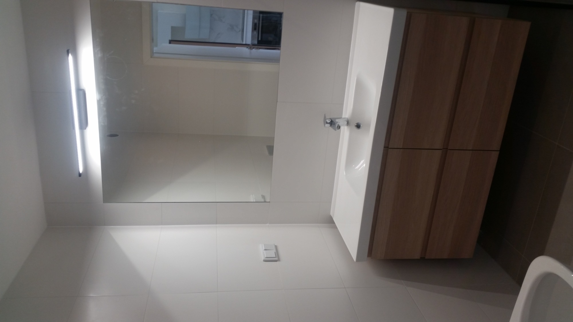 Nowoczesna łazienka z białymi płytkami, drewnianą szafką pod umywalką z białym blatem i minimalistycznym oświetleniem LED nad lustrem.