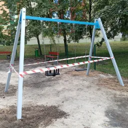 Plac zabaw z huśtawkami ogrodzony taśmą ostrzegawczą, widoczne ślady wykopów w piasku, w tle drzewa i budynek mieszkalny.