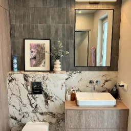 Nowoczesna łazienka z szarymi płytkami pionowymi, marmurowym wykończeniem, białą umywalką nablatową na drewnianej szafce, lustrem z podświetleniem LED oraz obrazem w czarnej ramie.