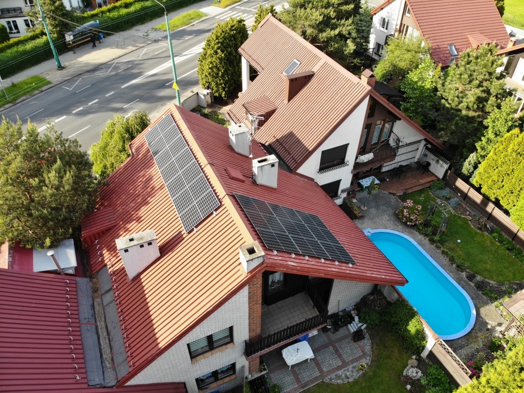 Widok z góry na dom z czerwoną dachówką i panelami fotowoltaicznymi, obok basen i zadbany ogród, w tle ulica i inne domy.