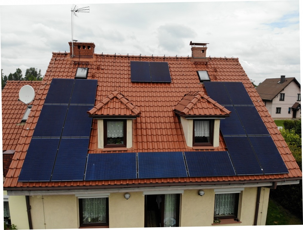 Instalacja paneli słonecznych na dachu domu z dachówką ceramiczną, widok z góry na panele, okna dachowe i komin. Na dachu antena telewizyjna.