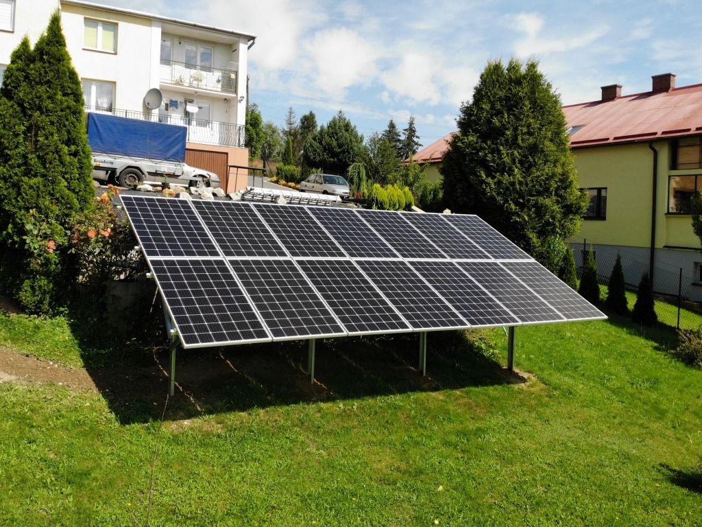 Panel słoneczny na metalowej konstrukcji, trawnik, w tle domy i drzewa w słoneczny dzień.