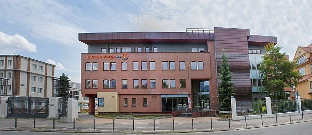 Panorama budynku Centrum Konferencyjnego z ceglaną fasadą, brązowymi elementami wykończeniowymi i pomarańczowym napisem na elewacji, otoczonego zielenią i ogrodzeniem.