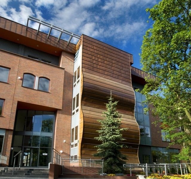 Nowoczesny budynek biurowy z fasadą z miedzianych paneli o falistym kształcie, drzewko iglaste przed wejściem, błękitne niebo w tle.