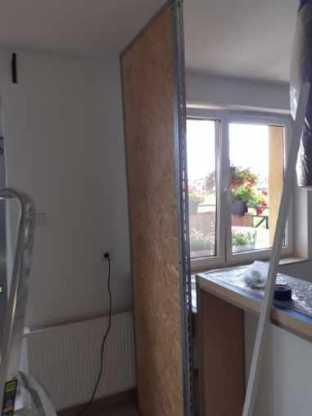 Płyta OSB z metalową ramą oparta o ścianę w pomieszczeniu w trakcie prac wykończeniowych, widoczny fragment drabiny i okno z kwiatami.