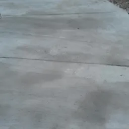 Świeżo wylany, szary betonowy podjazd z widocznymi śladami wilgoci i szczelinami dylatacyjnymi, graniczący z ziemią i fragmentem ciemnej ściany.