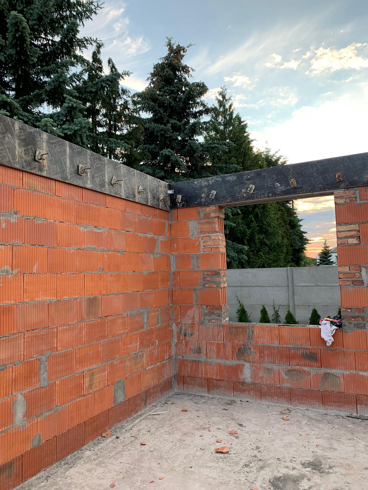 Surowa ściana z czerwonej cegły ceramicznej w trakcie budowy, z widocznymi elementami szalunku i kotwami, na tle zielonych drzew i błękitnego nieba o zmierzchu.