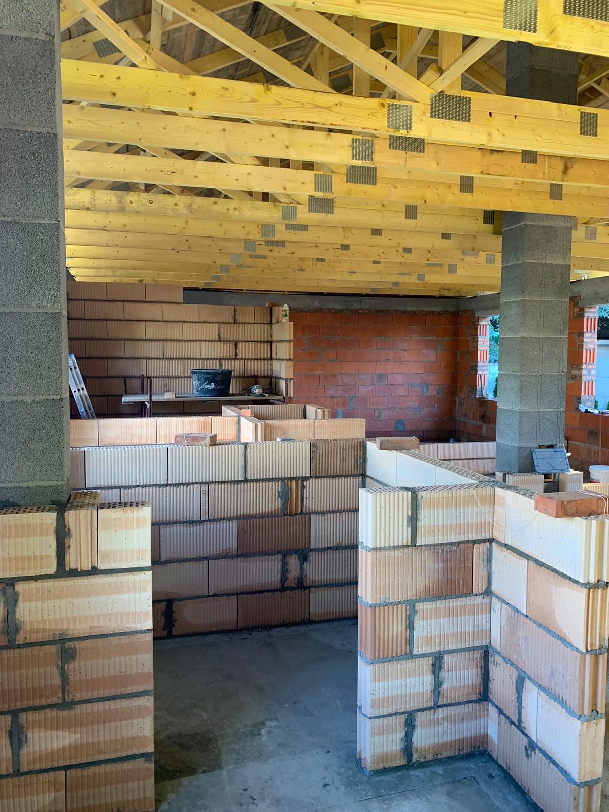 Wnętrze w trakcie budowy: ściany z pustaków ceramicznych, drewniana konstrukcja dachu z metalowymi łącznikami, widoczne elementy murarskie i betonowe filary.