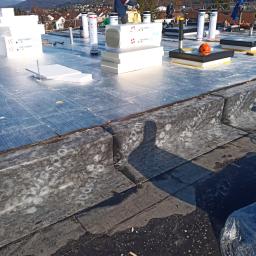 Dr. Dach Walkowiak - Prace na dachu płaskim w słoneczny dzień: dekarz pracuje nad izolacją, widoczne płyty izolacyjne Swissopor, narzędzia, oraz obróbki blacharskie. W tle zabudowania mieszkalne i zalesione wzgórza.