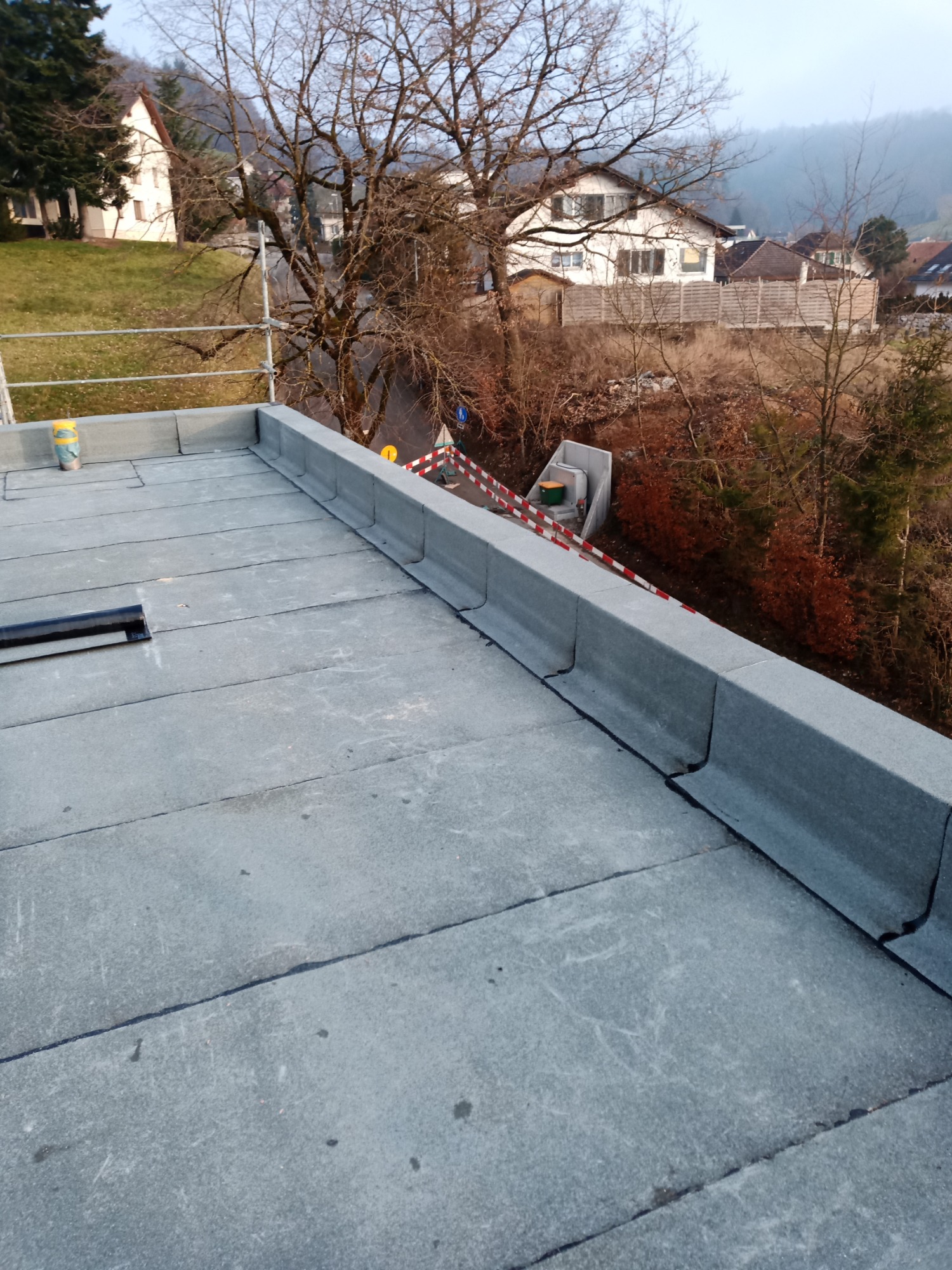 Szary płaski dach z papy termozgrzewalnej z obróbką blacharską na krawędzi, widok z góry na tle zabudowań mieszkalnych i drzew bez liści.