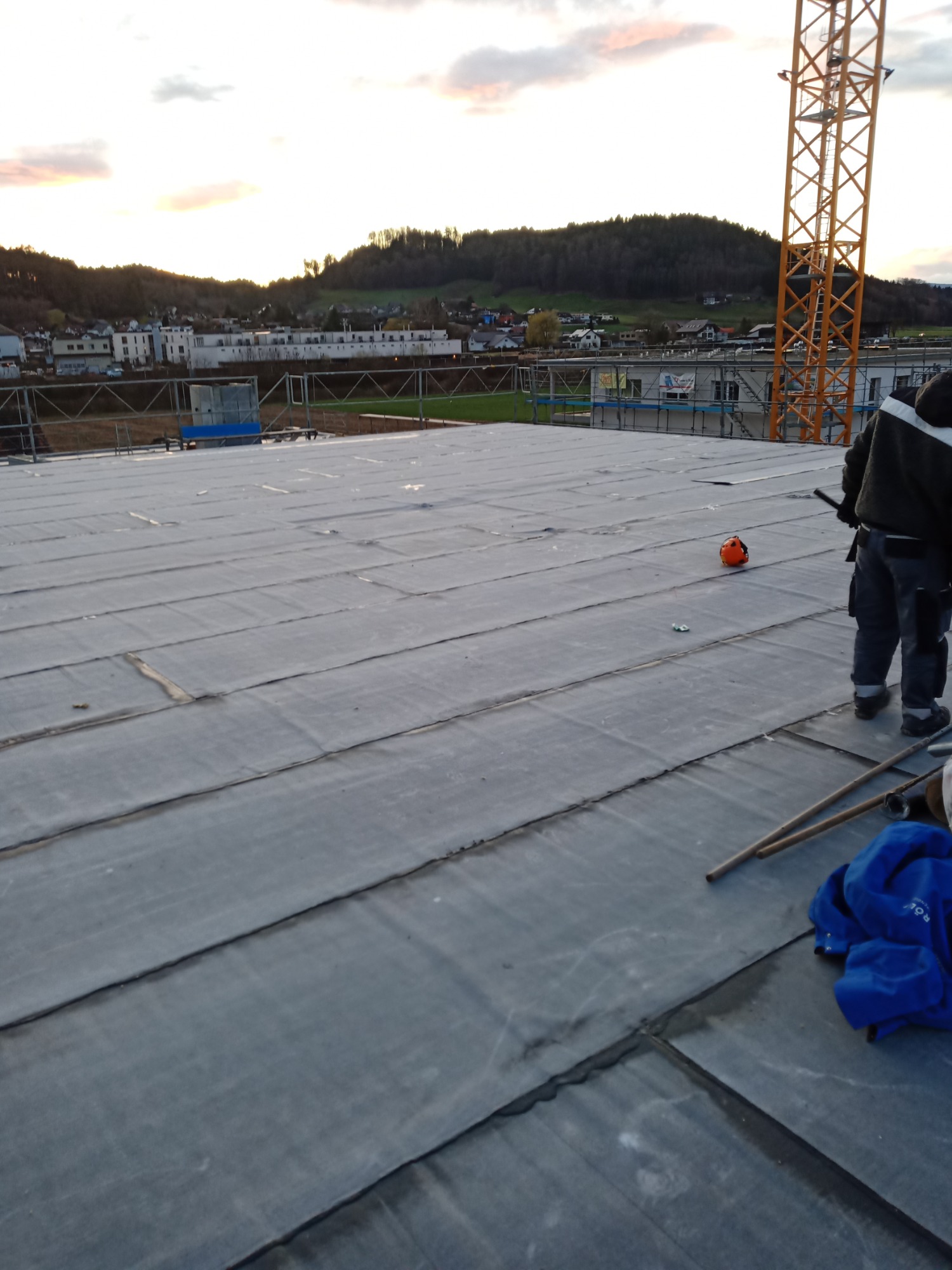 Widok z góry na płaski dach pokryty szarą papą, z elementami konstrukcyjnymi, pomarańczowym kaskiem i pracownikiem w tle oraz żurawiem budowlanym i zabudową w oddali, w tle zachodzące słońce.