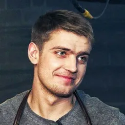 Uśmiechnięty młody mężczyzna w szarej koszulce i skórzanym fartuchu, z krótkimi blond włosami i lekkim zarostem, spoglądający w bok na tle ciemnej ściany.