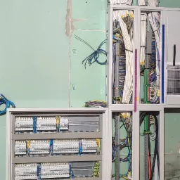 Rozdzielnica elektryczna z licznymi przewodami i zabezpieczeniami, zamontowana na ścianie z płyt gipsowo-kartonowych, widoczne koryta kablowe pod sufitem.