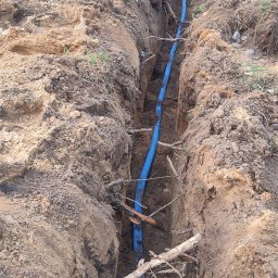 Smart Home Leszno - Inteligentny Dom IGVEL - Głogów, Rawicz, Gostyń, Kościan, Śrem - Niebieski przewód karbowany ułożony w wykopie ziemnym, widoczne gałęzie. Etap prac instalacyjnych, teren budowy.