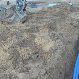 Smart Home Leszno - Inteligentny Dom IGVEL - Głogów, Rawicz, Gostyń, Kościan, Śrem - Wykop z ułożonymi niebieskimi rurami kablowymi, obok koparka E19. Widoczne etapy prac ziemnych pod instalacje elektryczne lub teletechniczne.