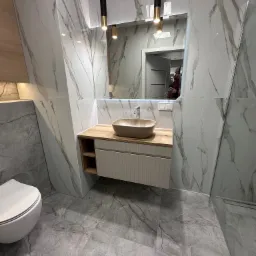 Nowoczesna łazienka z marmurowymi płytkami, beżową umywalką nablatową i wiszącą toaletą. Widoczne oświetlenie punktowe i fragment kabiny prysznicowej.