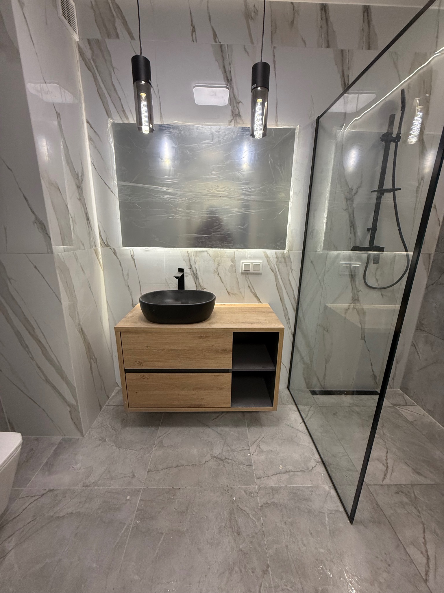 Nowoczesna łazienka z marmurowymi płytkami, drewnianą szafką pod umywalką z czarną misą i kabiną prysznicową z czarnym obramowaniem.