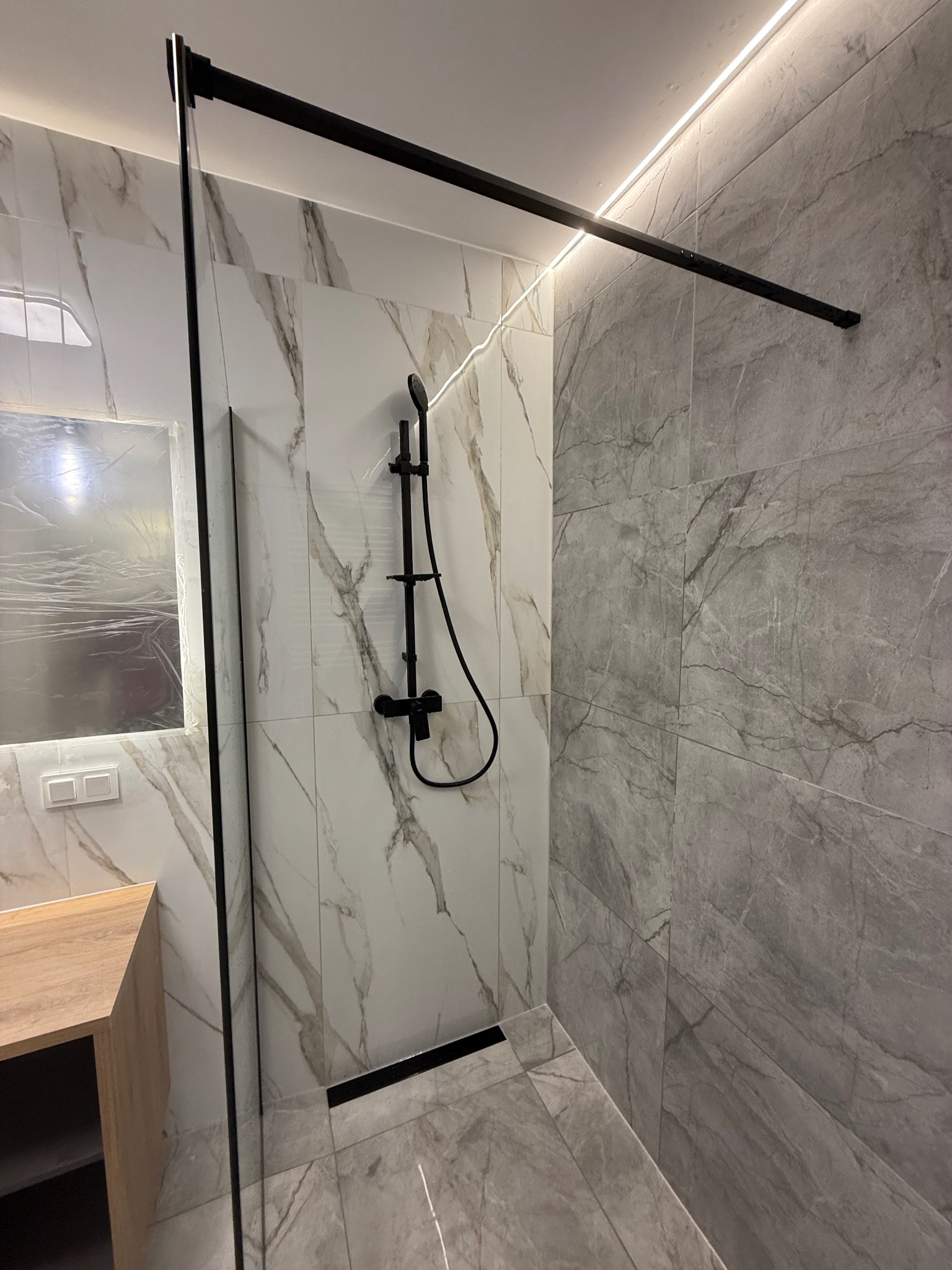 Nowoczesna kabina prysznicowa z czarną armaturą na tle marmurowych płytek w odcieniach szarości i bieli, oświetlona listwą LED.