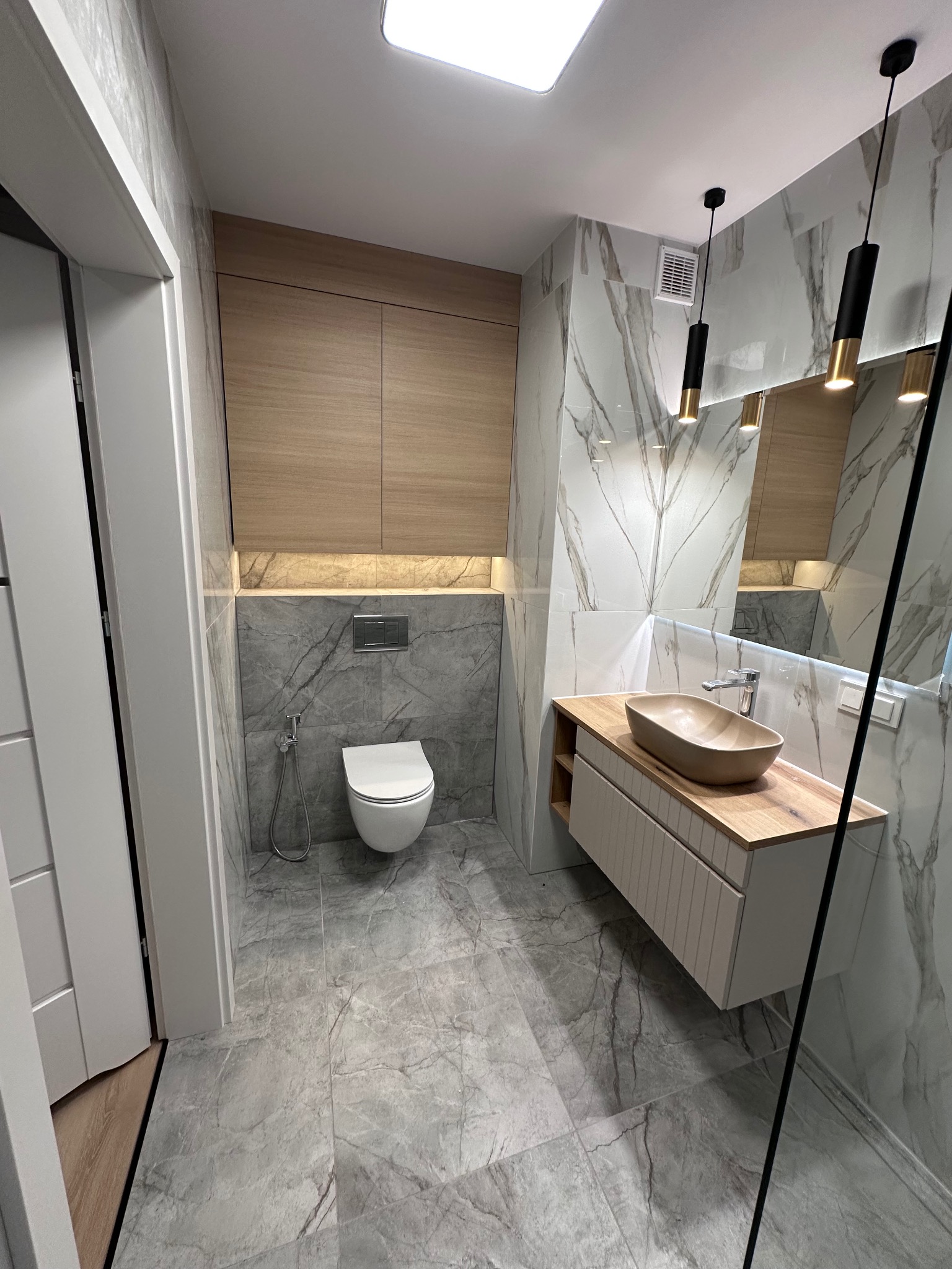 Nowoczesna łazienka z marmurowymi płytkami, drewnianymi szafkami i umywalką w kształcie misy. Czarne, wiszące lampy dodają elegancji. Szare płytki podłogowe.
