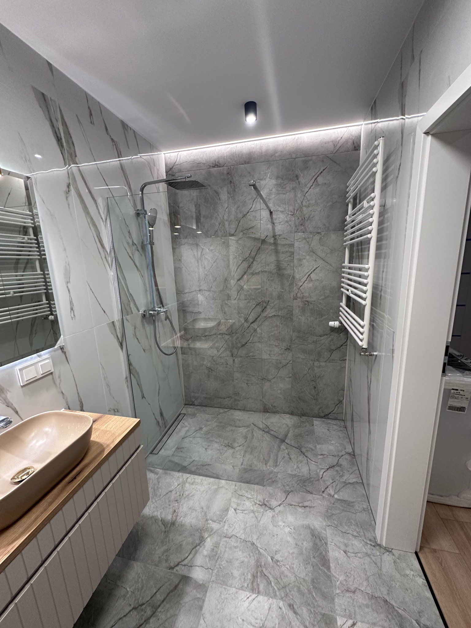 Nowoczesna łazienka z kabiną prysznicową typu walk-in, marmurowymi płytkami i oświetleniem LED. Umywalka nablatowa w kolorze beżowym.