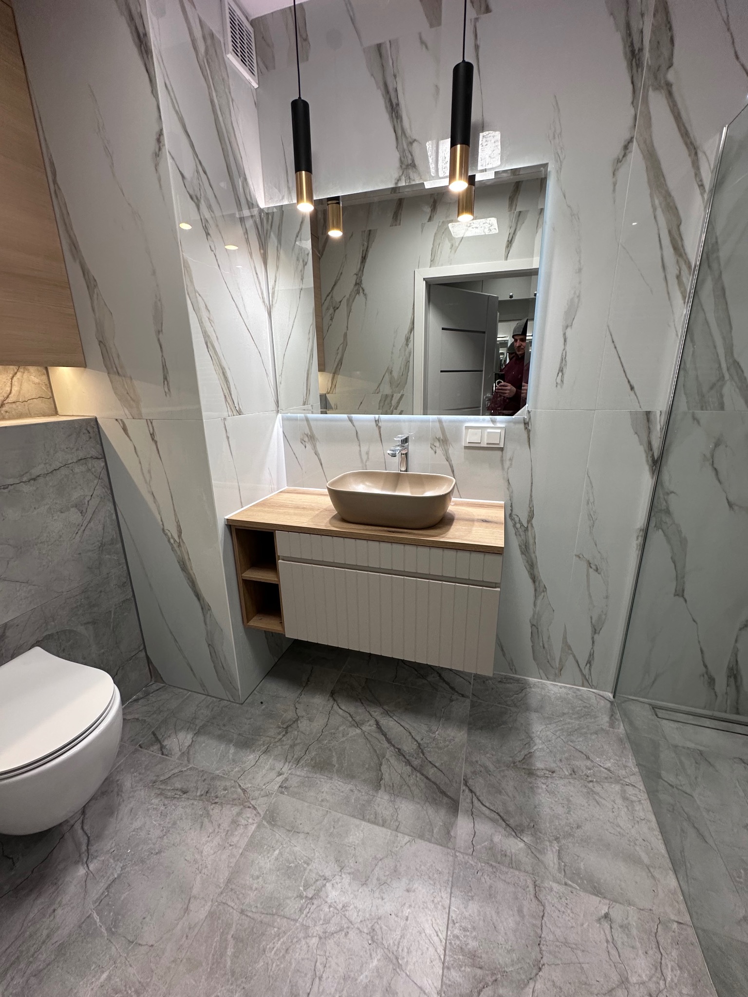 Nowoczesna łazienka z marmurowymi płytkami, beżową umywalką nablatową i wiszącą toaletą. Widoczne oświetlenie punktowe i fragment kabiny prysznicowej.