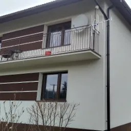 Dwukondygnacyjny dom z ocieploną, białą elewacją i brązowymi akcentami, widoczne balkony z metalowymi balustradami, antena satelitarna i pracownik na rusztowaniu przy oknie.