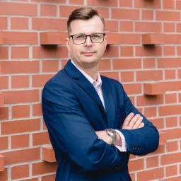 Portret mężczyzny w okularach i granatowym garniturze, stojącego na tle ceglanej ściany z założonymi rękami i srebrnym zegarkiem na nadgarstku.