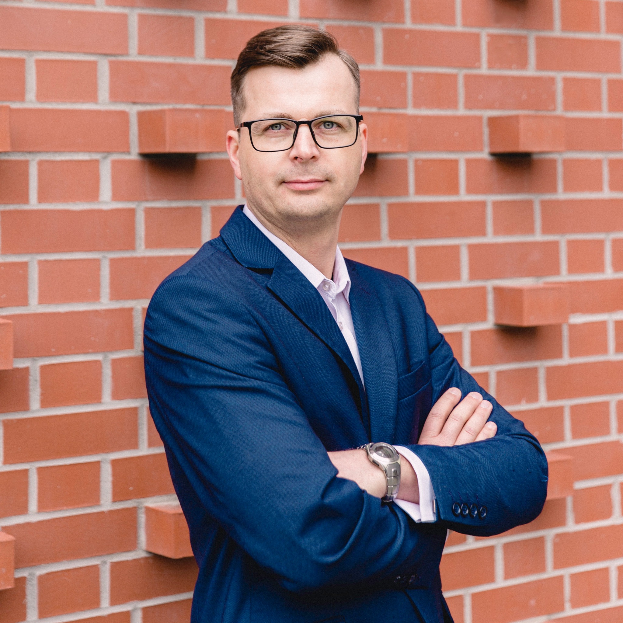Portret mężczyzny w okularach i granatowym garniturze, stojącego na tle ceglanej ściany z założonymi rękami i srebrnym zegarkiem na nadgarstku.