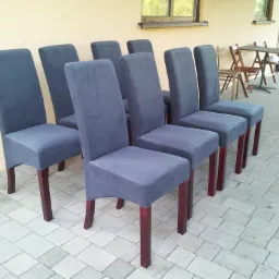 Rząd ośmiu krzeseł z ciemnoszarą tapicerką i mahoniowymi nogami, ustawionych na brukowanym patio przed budynkiem z oknami.