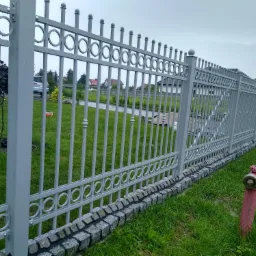 Szare metalowe ogrodzenie z dekoracyjnymi okręgami i kamienną podmurówką, widok na posesję z domem i hydrant w trawie.