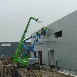 Montaż paneli warstwowych na hali przemysłowej z użyciem podnośników koszowych; trzech pracowników w kaskach i kamizelkach odblaskowych na wysokości, jeden z nich macha ręką; w tle widoczny budynek...