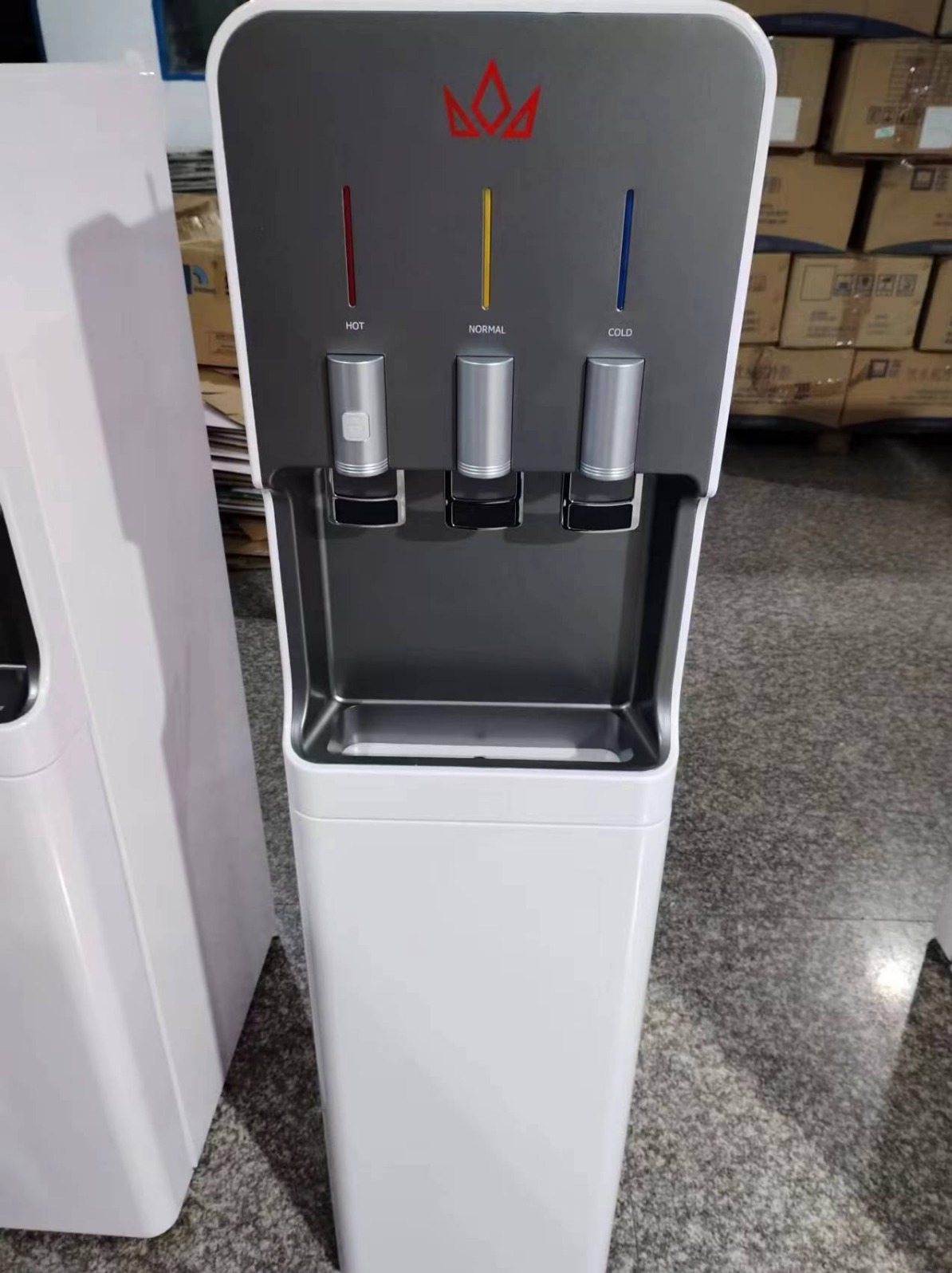 Dystrybutor wody z trzema kranami: gorąca, normalna i zimna, z podświetleniem w kolorach czerwonym, żółtym i niebieskim, widoczny w tle magazyn z kartonami.