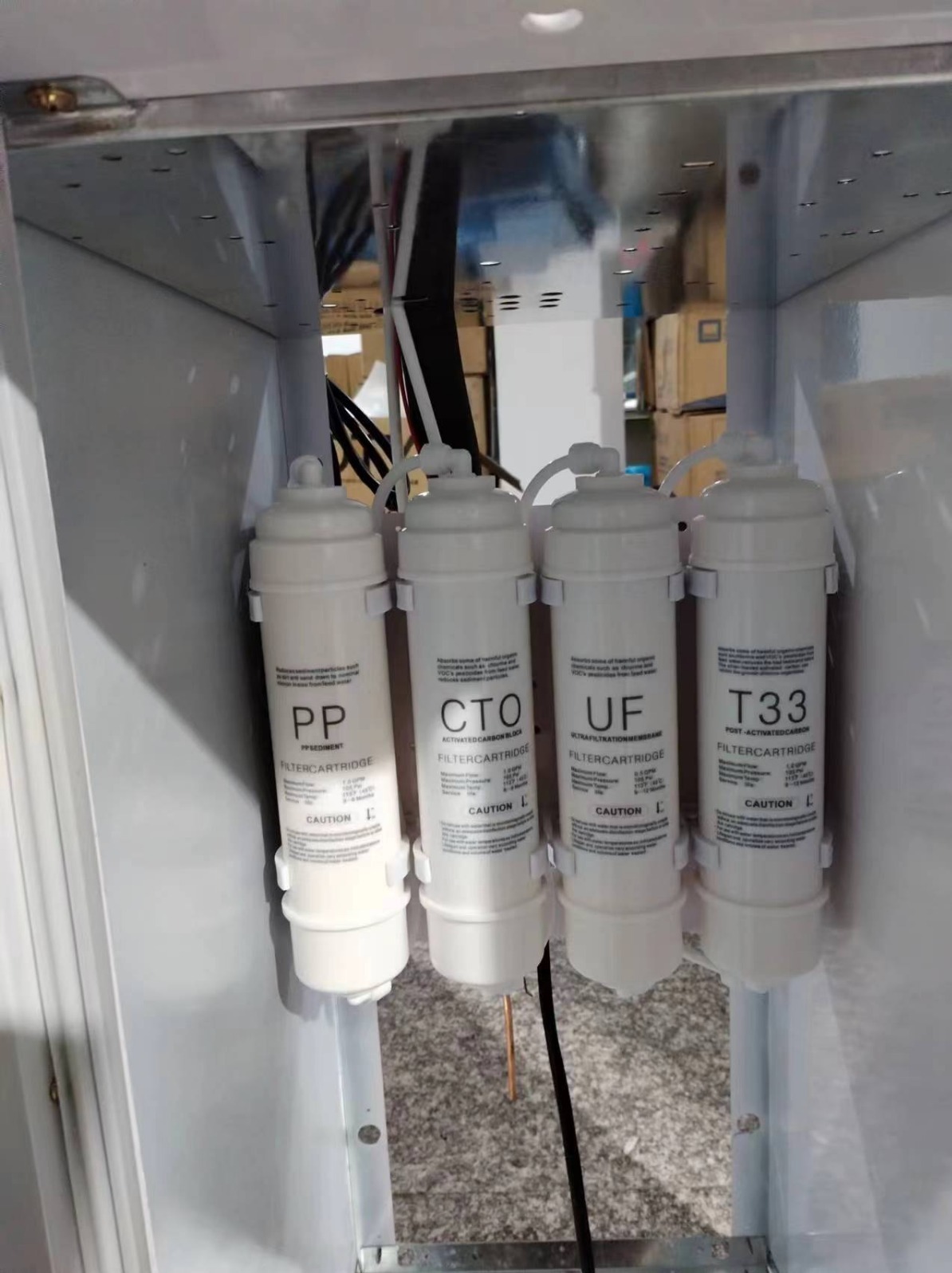 Wnętrze urządzenia do uzdatniania wody z czterema cylindrycznymi filtrami: PP, CTO, UF, T33, każdy z etykietą i ostrzeżeniem 'Caution'.