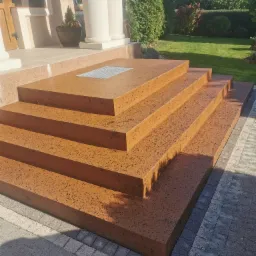 Zewnętrzne schody z posadzki żywicznej w kolorze terakoty, prowadzące do wejścia z kolumnami, z kratką odpływową na najwyższym stopniu i kostką brukową u podstawy.