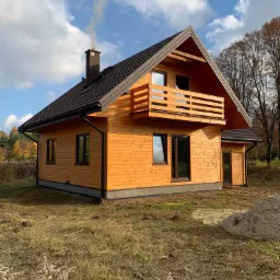 Nowoczesny dom szkieletowy z drewnianą elewacją i balkonem, z komina unosi się dym, otoczony jesiennym krajobrazem.