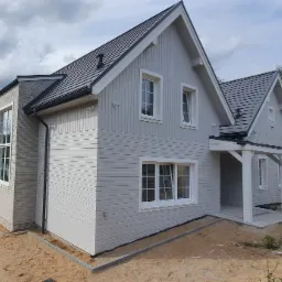 Jasnoszary dom z drewnianą elewacją i ciemnoszarą dachówką, z widocznym fragmentem dobudówki z cegły, w otoczeniu piaszczystego terenu i drzew.