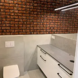 Nowoczesna łazienka z umywalką nablatową na białej szafce z czarnymi uchwytami, toaleta podwieszana i ścianą wykończoną czerwoną cegłą.